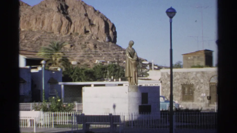 Guaymas 8mm travel frame from 1975 with a statue on a white base, nearby streetlamp, fenced park, palm trees, buildings, and rocky hillside.