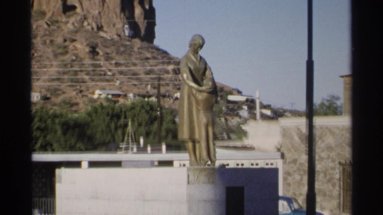 Mother-and-child statue centered between lampposts with hillside steps and a parked blue car nearby.