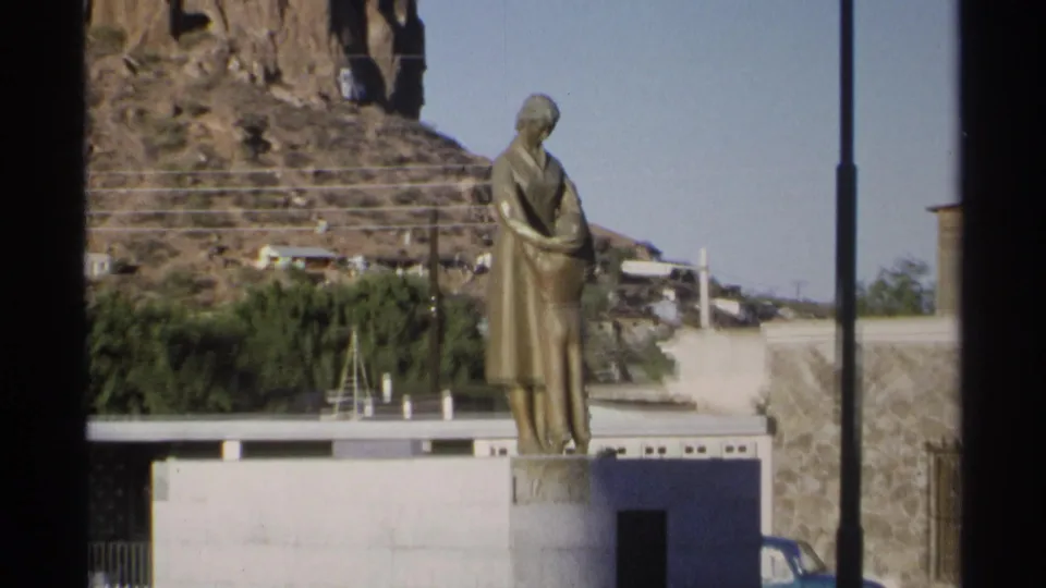 1975 Guaymas frame with the bronze mother-and-child statue centered in a plaza, hillside and blue car visible behind.