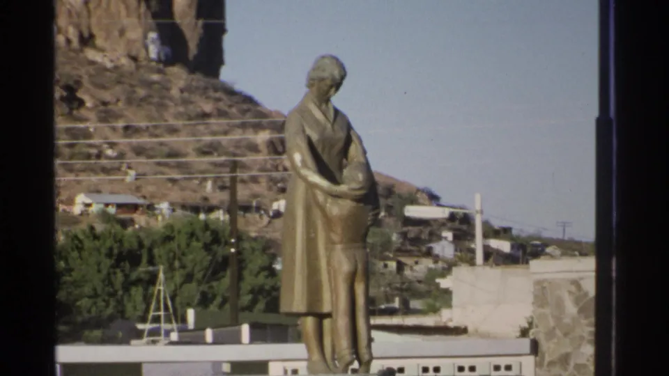 Closer 1975 Guaymas 8mm view of the bronze mother-and-child sculpture against hillside homes and trees.