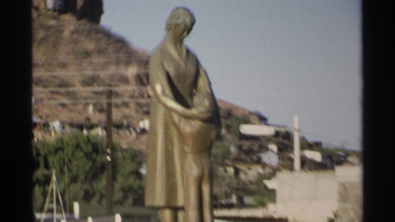Bronze statue of a woman holding a child with a hillside and houses behind it.