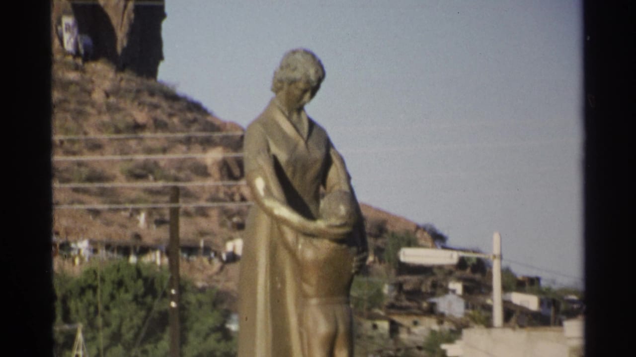 Bronze mother-and-child statue beside a hillside with houses and power lines.