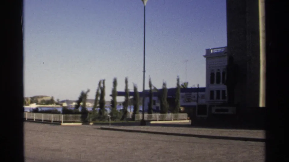 1975 Guaymas 8mm frame showing a paved waterfront plaza, cypress trees, a white building, and the sea in the distance.