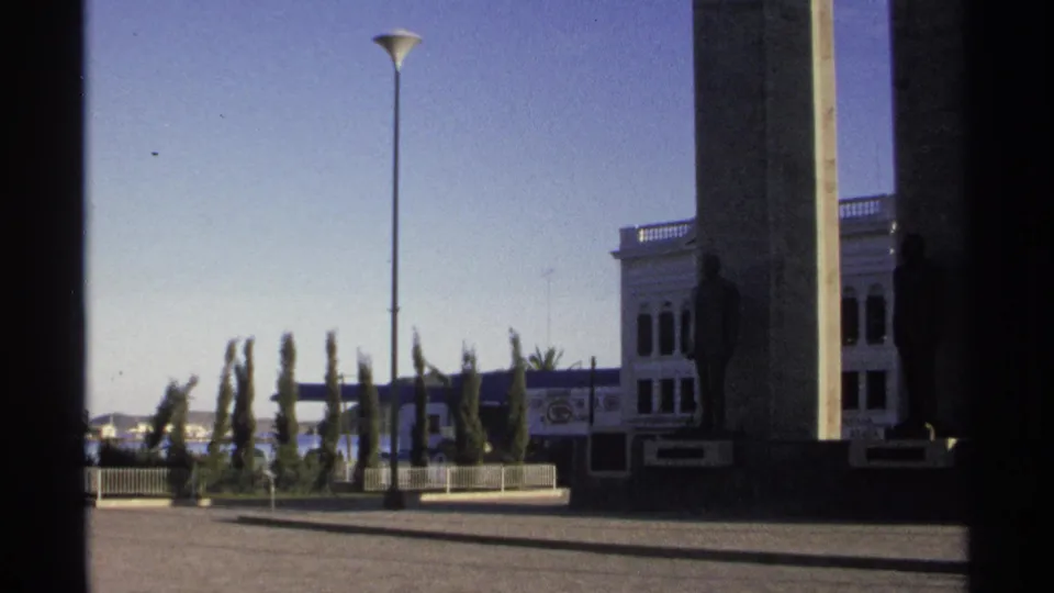 1975 Guaymas home-movie frame of a waterfront plaza with a tall lamppost, cypress trees, building columns, and distant sea.