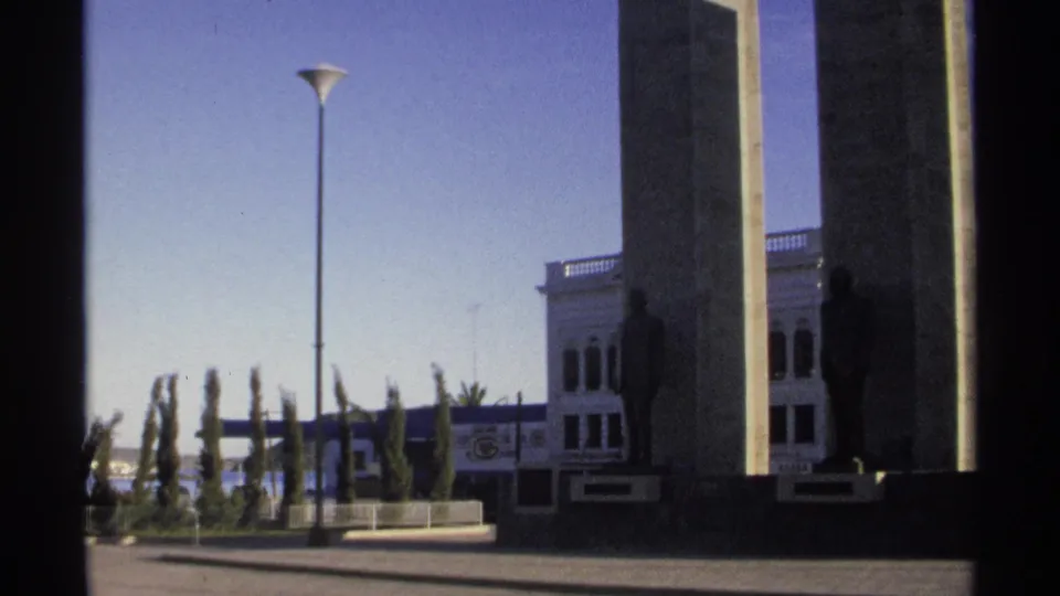 1975 Guaymas 8mm view of a civic monument with tall concrete pillars, bronze statues, a street lamp, and blue sky.