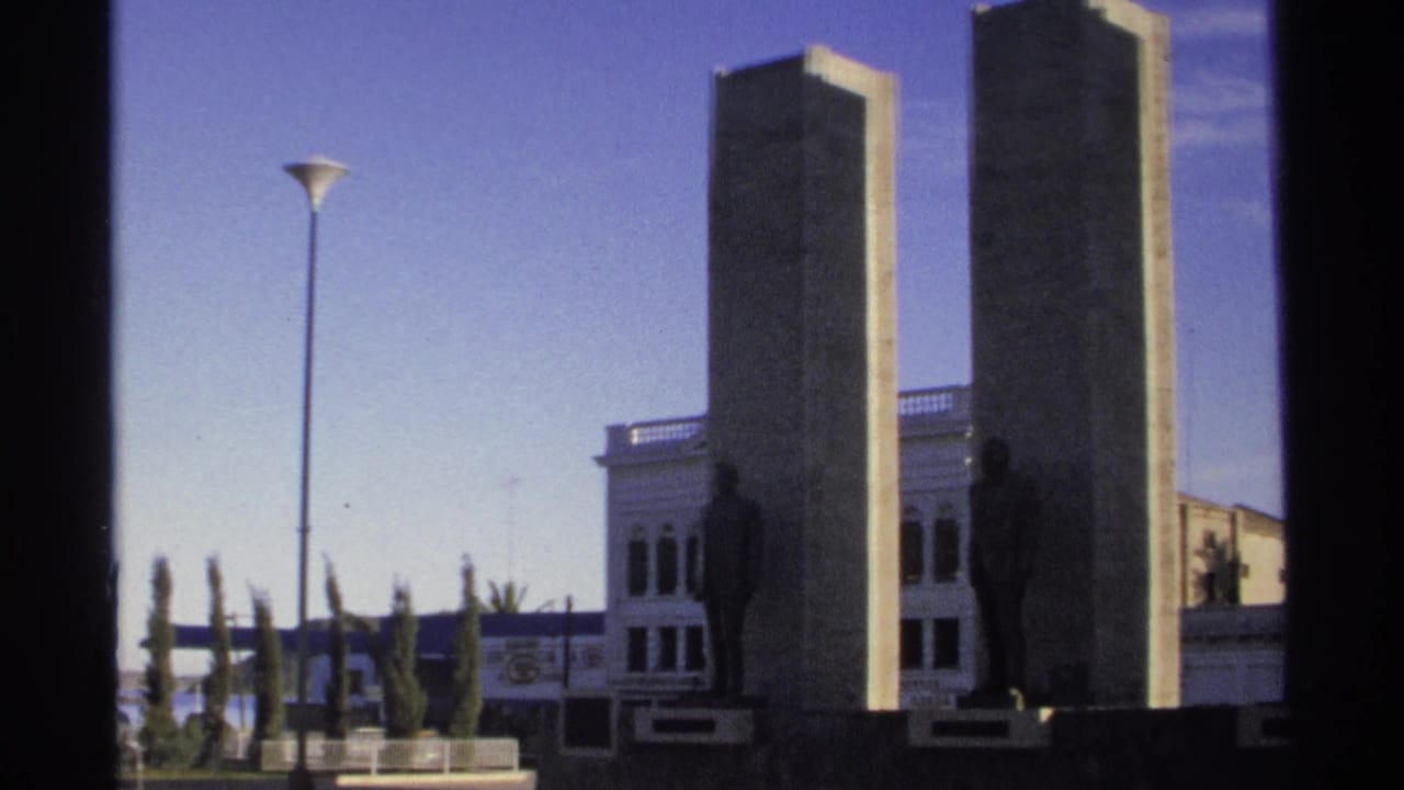 Concrete monument pillars with bronze figures beside a white building.