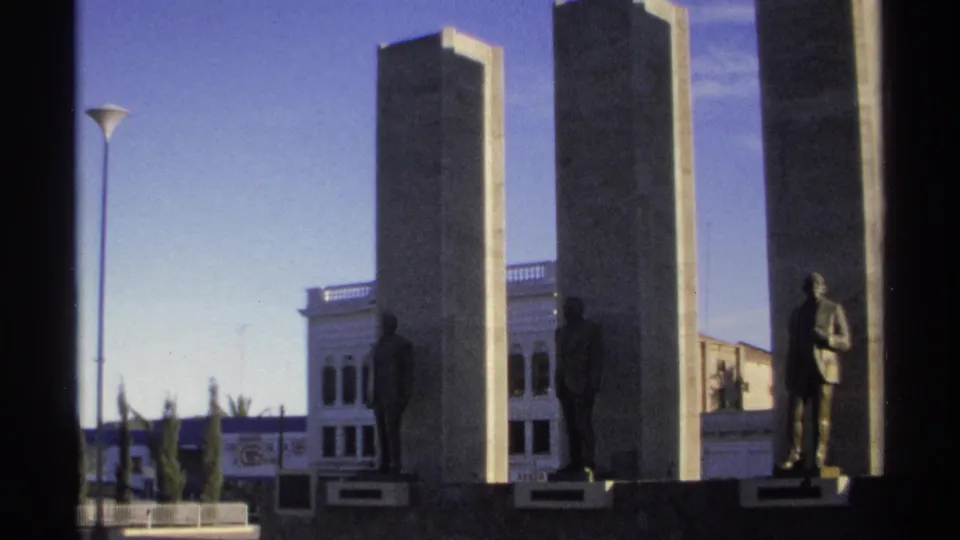 A wider 1975 Guaymas 8mm frame shows the monument, bronze statues, street lamp, plantings, and adjacent urban buildings.