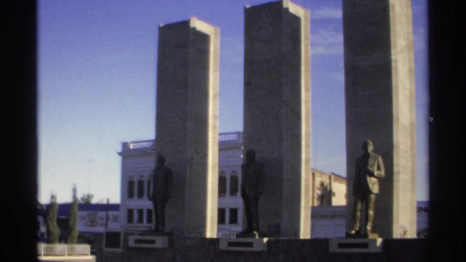 1975 Guaymas home-movie frame of sunlit concrete pillars with bronze suited figures and a white building in the background.