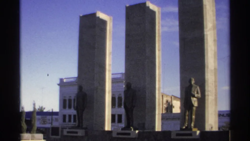 Guaymas, Mexico home-movie frame from 1975 showing a civic monument with tall stone pillars and bronze standing statues before a white building.