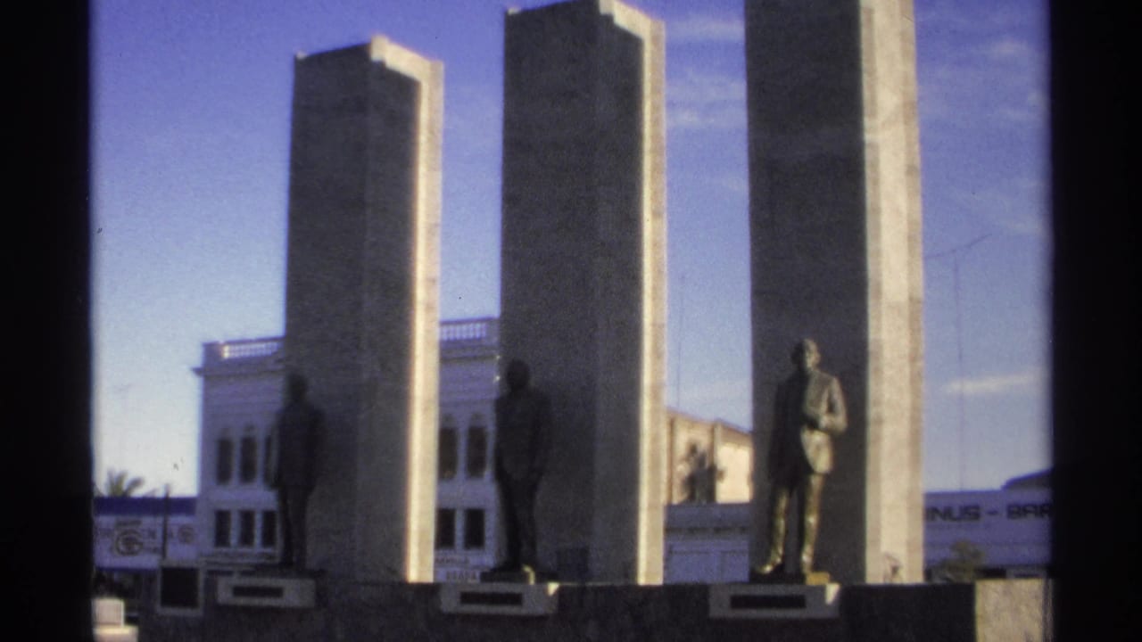 Three tall pillars and statues seen across a street under a blue sky.