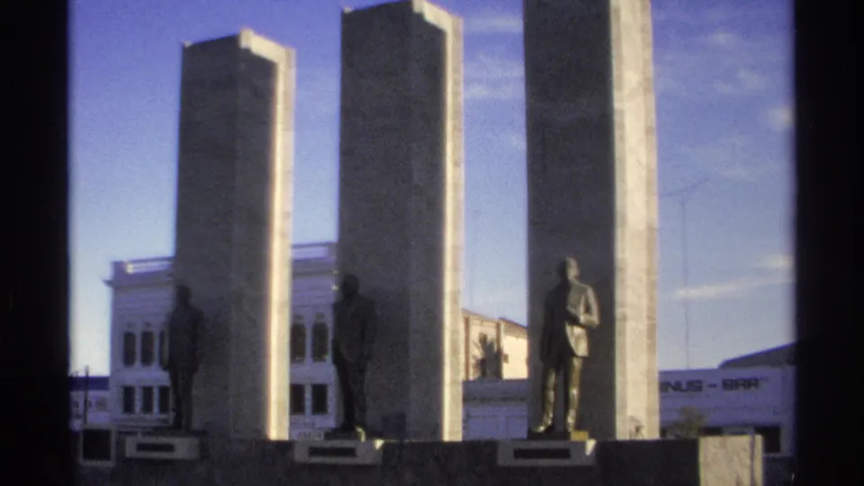 A closer 1975 Guaymas frame shows the stone monument, standing statues on plinths, and the white building behind it, with partial signage at the edge.
