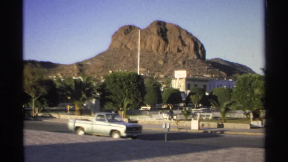 A light-colored pickup truck moves through a Guaymas street scene with trimmed trees, signage, low buildings, and a rocky hill rising behind the town.