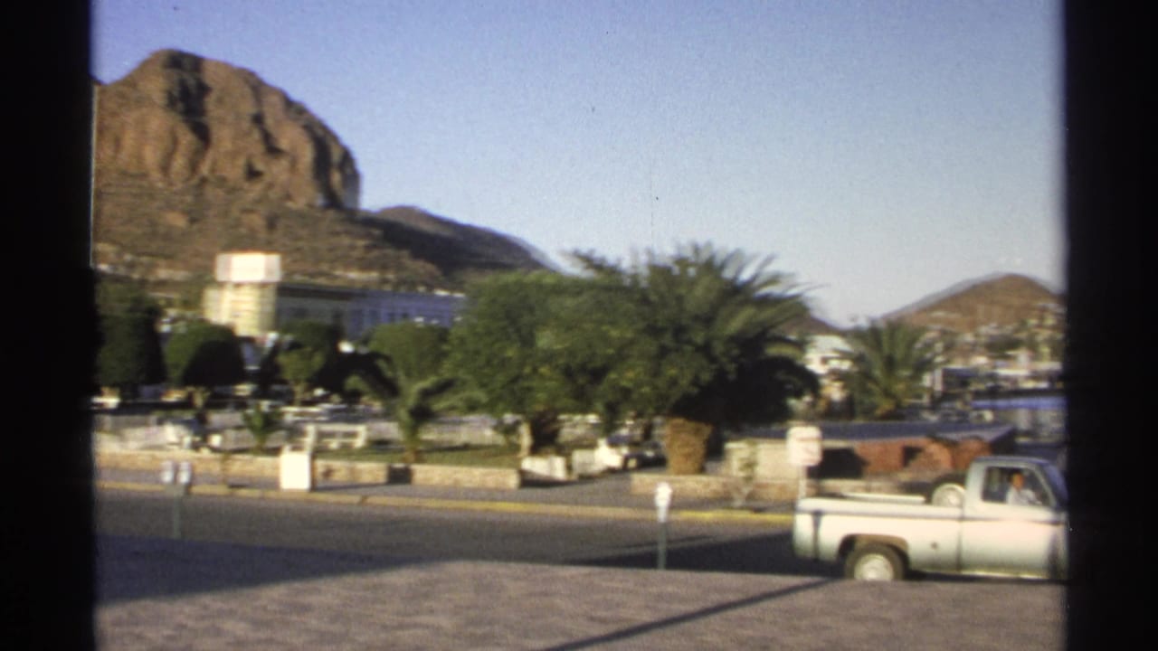 Guaymas, Mexico street scene with palms, vehicles, a rocky hill, and distant waterfront glimpsed through window framing.