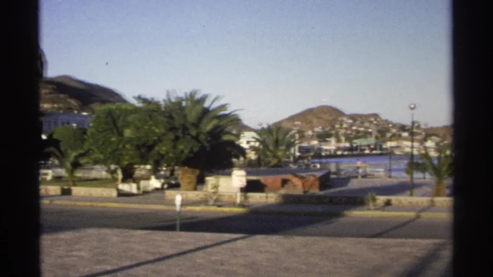1975 home-movie view of a Guaymas street, landscaped palms, blue water, and hillside buildings beyond the road.