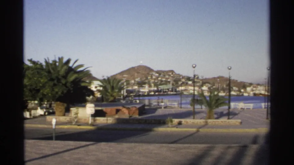 Guaymas waterfront view with palms, a paved road, hillside buildings, and blue water framed by dark 8mm film edges.