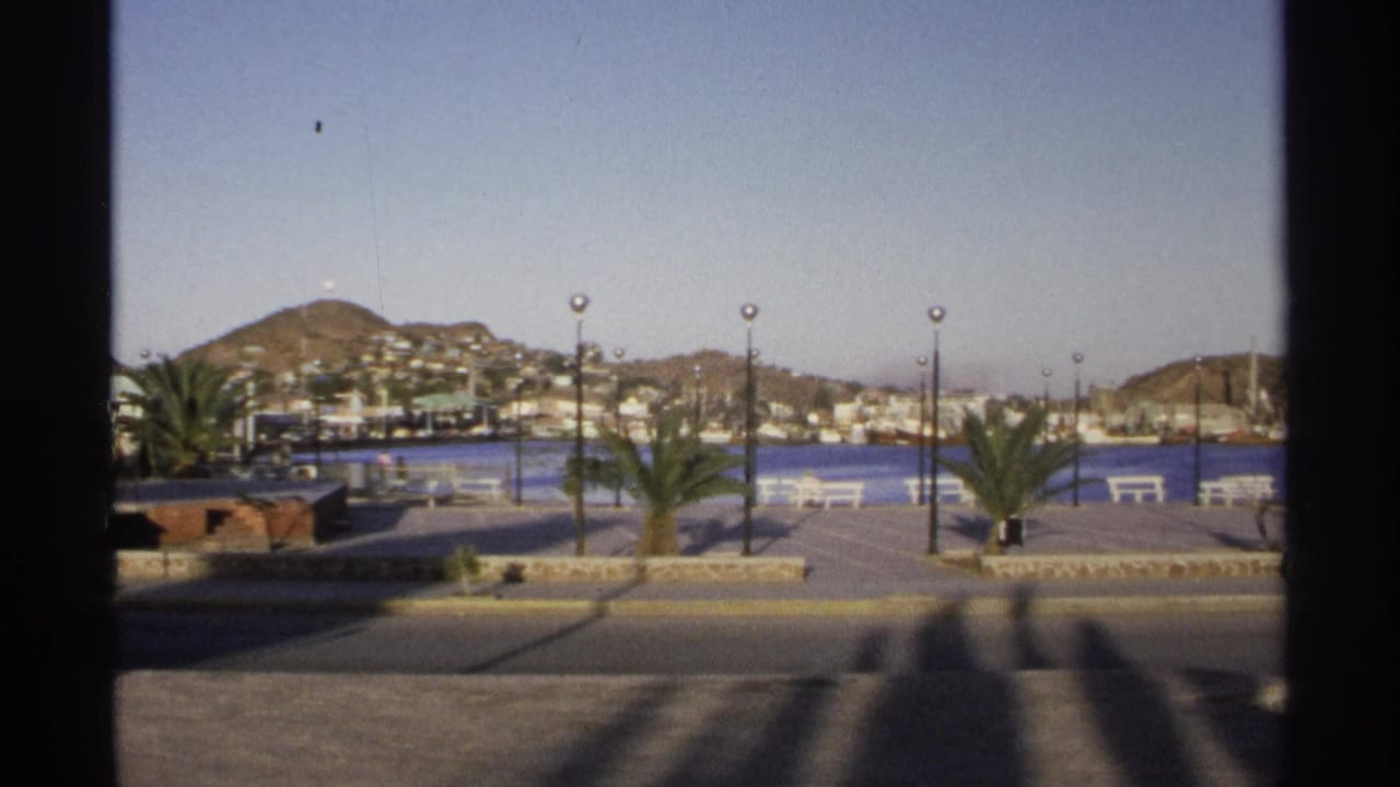 Promenade beside blue harbor water with palms, benches, lampposts, boats, hillside buildings, and mountains.