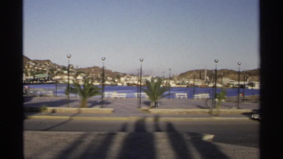 1975 8mm frame of the Guaymas waterfront, with a paved promenade, palms, white benches, lampposts, blue harbor water, hillside buildings, and a street in the foreground.