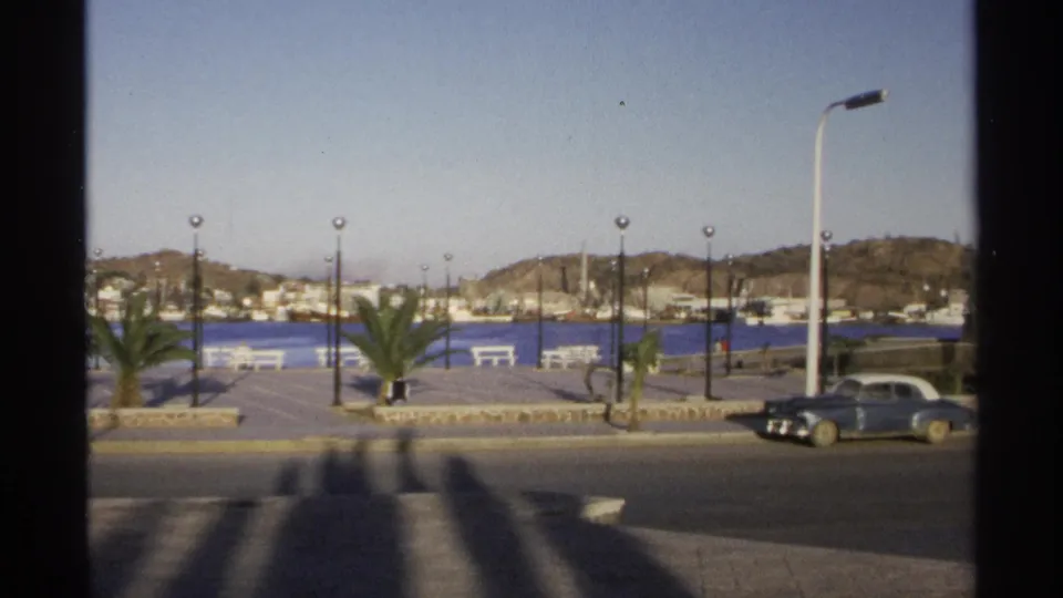 1975 Guaymas street and waterfront view with a blue older car at the curb, palm-lined promenade, white benches, lampposts, harbor water, hillside buildings, and mountains.