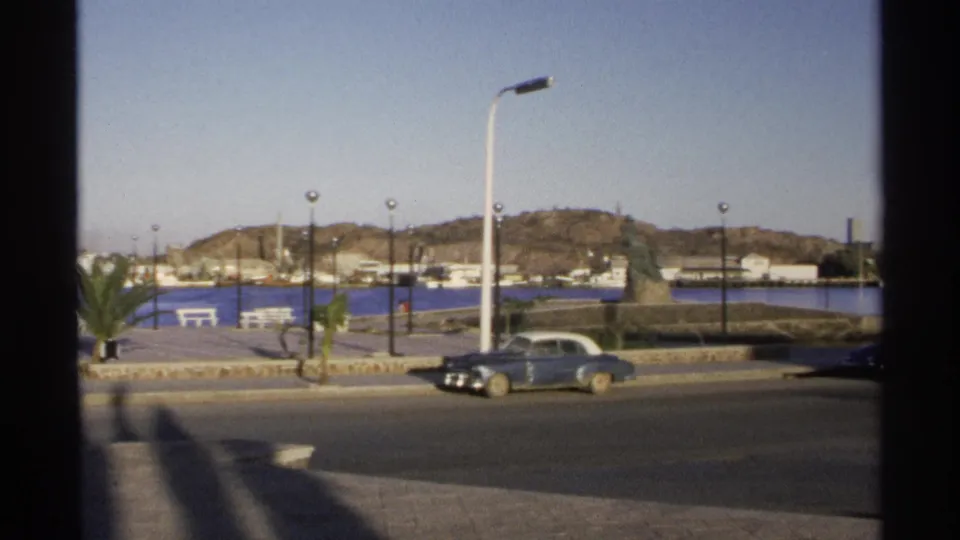Guaymas, Mexico harbor road with classic sedan beside a waterfront monument, promenade lamps, palm plantings, and boats in the distance.