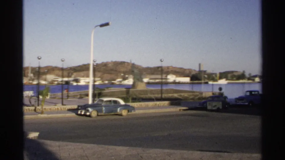 1975 Guaymas street scene showing a classic sedan, another parked or passing vehicle, waterfront monument, harbor water, lamps, and hillside.
