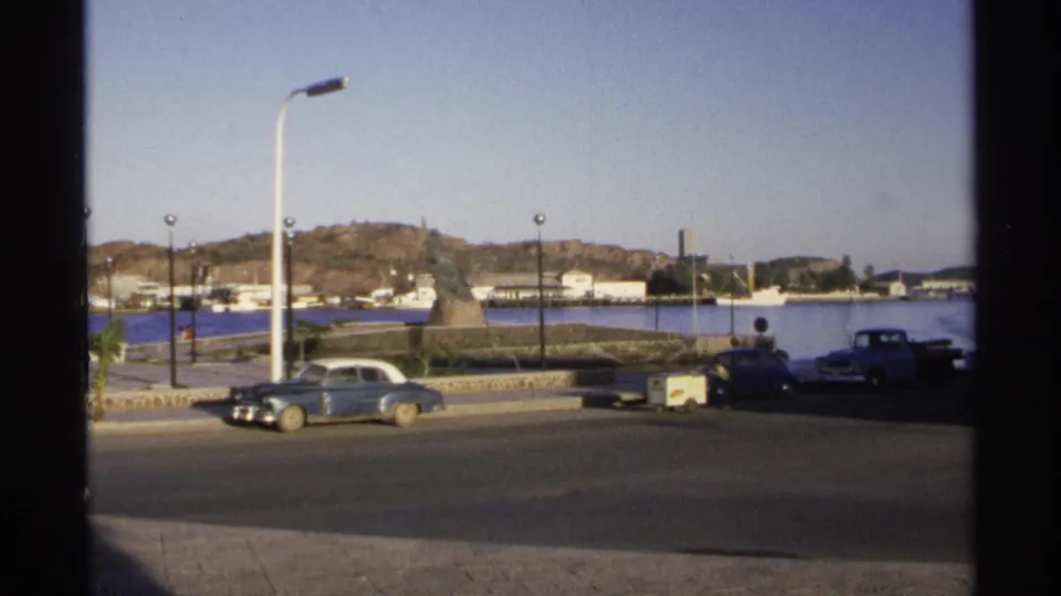 Guaymas waterfront view with classic cars near a monument, promenade lamps, harbor water, small boats, and a sunlit hillside.