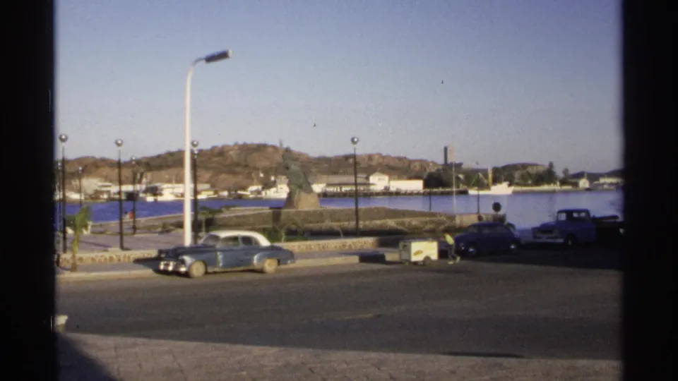 1975 Guaymas waterfront view with parked cars, calm harbor water, low hills, streetlights, and a monument near the road.