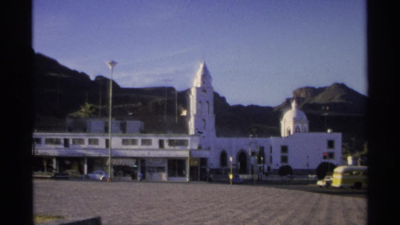 White church buildings beside a plaza with mountains behind and a few vehicles near the street.