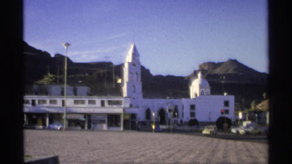 1975 Guaymas plaza view showing white church architecture, a domed chapel, a mountain backdrop, streetlights, and traffic at the edge.