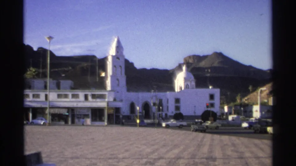 1975 Guaymas street and plaza view with white church buildings, vintage cars, scattered pedestrians, streetlights, and mountains beyond.