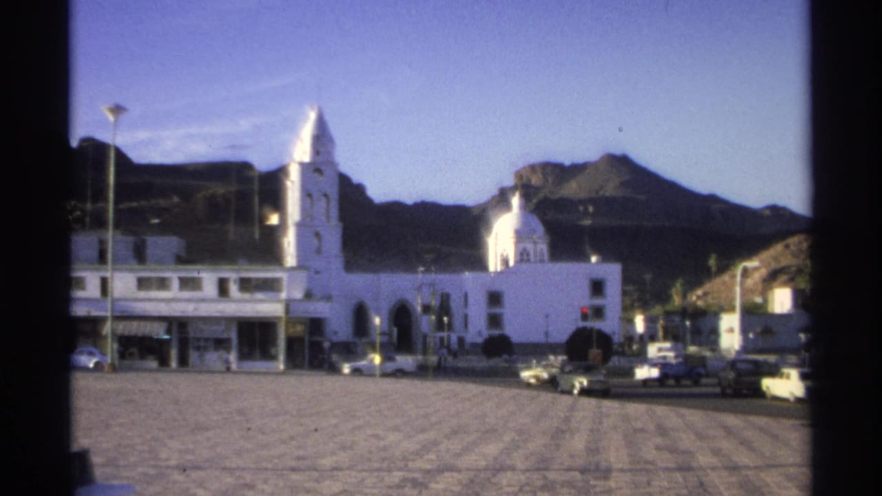 White church and bell tower across a paved town square with mountains behind.