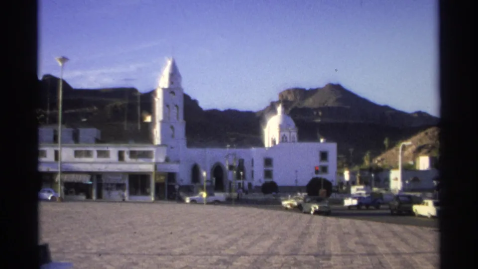 1975 Guaymas town square view with a white church, domed roof, parked cars, streetlamps, and dark mountains in the background.