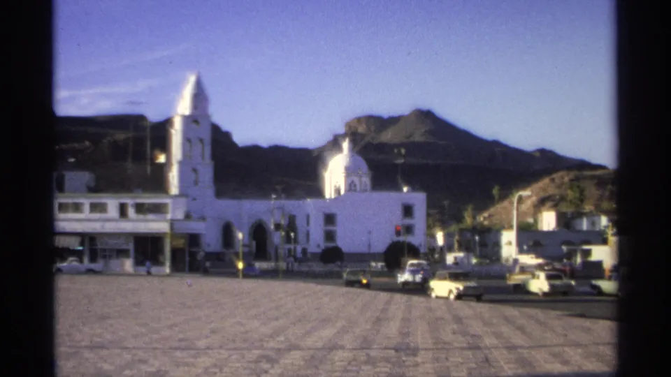 A white church and mountain ridge in Guaymas, Mexico, with 1970s cars parked and moving along the edge of the plaza.