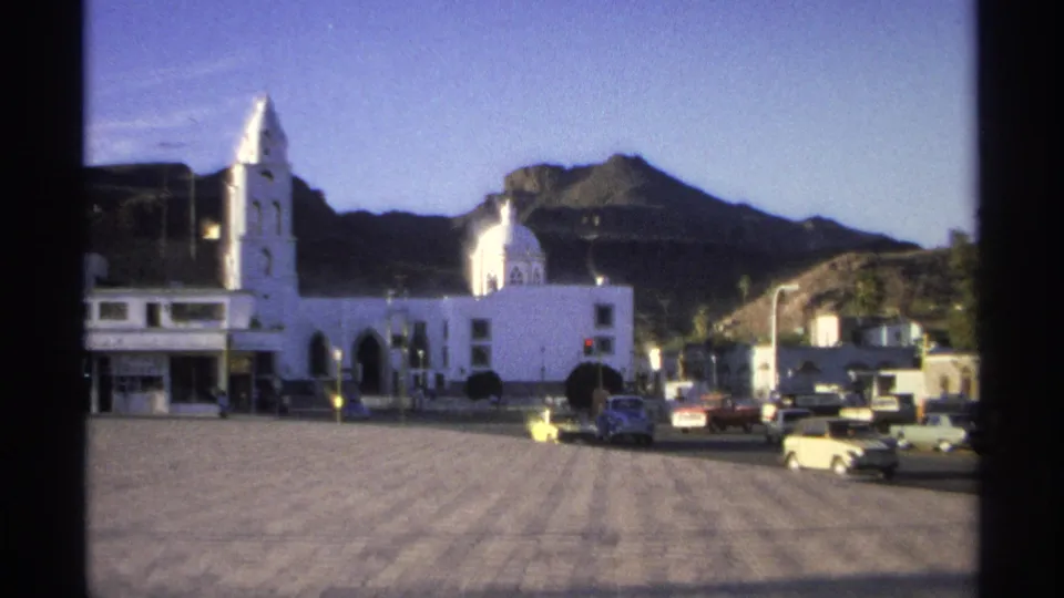 Guaymas plaza scene from 1975 showing the church tower, domed structure, passing cars, low buildings, and rugged hills beyond.