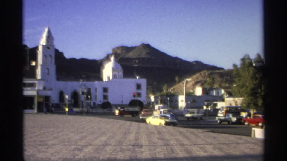 Late shot of the 1975 Guaymas square with a yellow sedan, other cars, the white church, trees, and mountains in warm light.