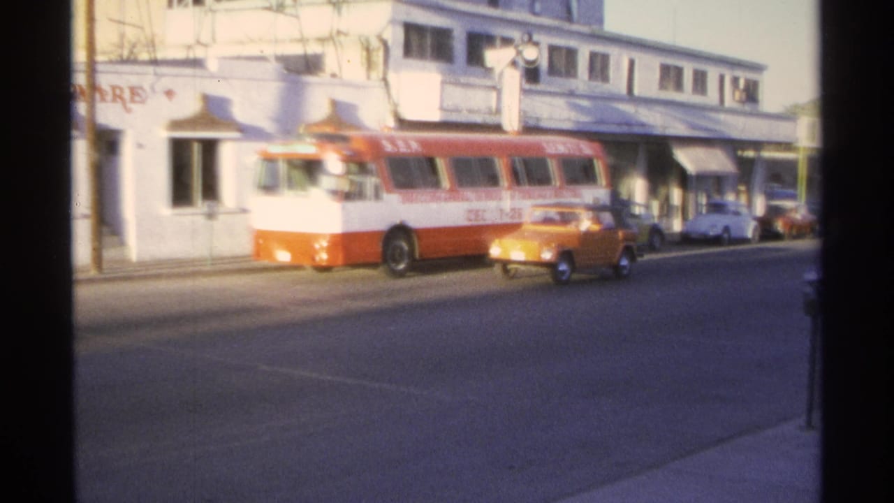 1970s Guaymas street view with a red-and-white bus, orange compact car, white storefront building, and parked cars.