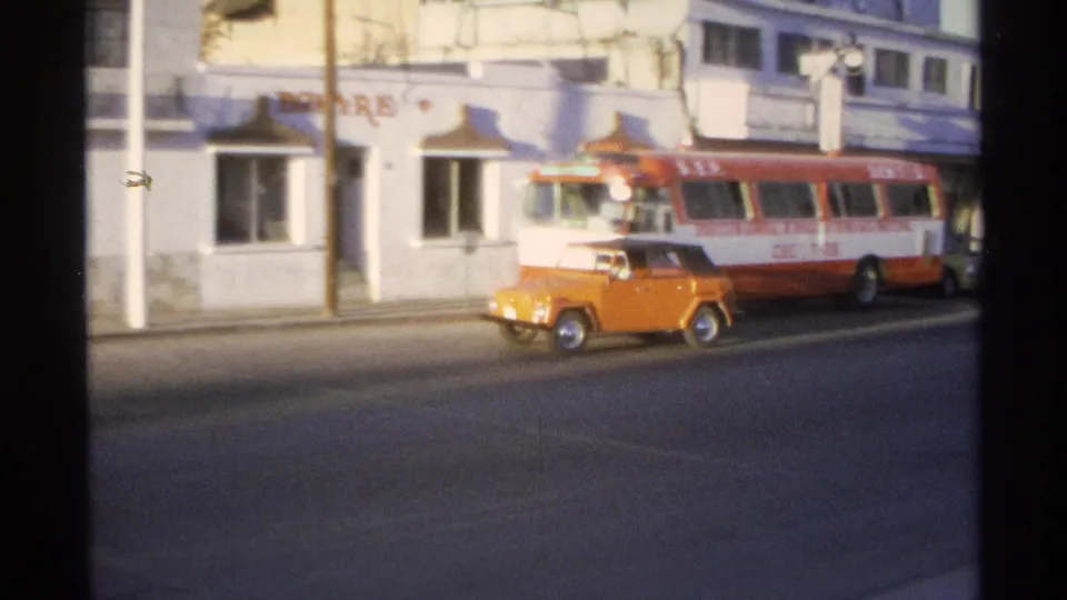 Orange compact car and red-and-white bus share a Guaymas street lined by a white commercial building.