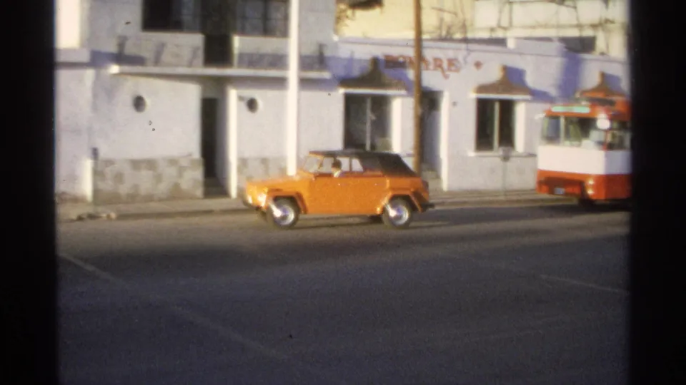 Small orange car on a Guaymas street beside a pale building, with the red-and-white bus partly entering the frame.