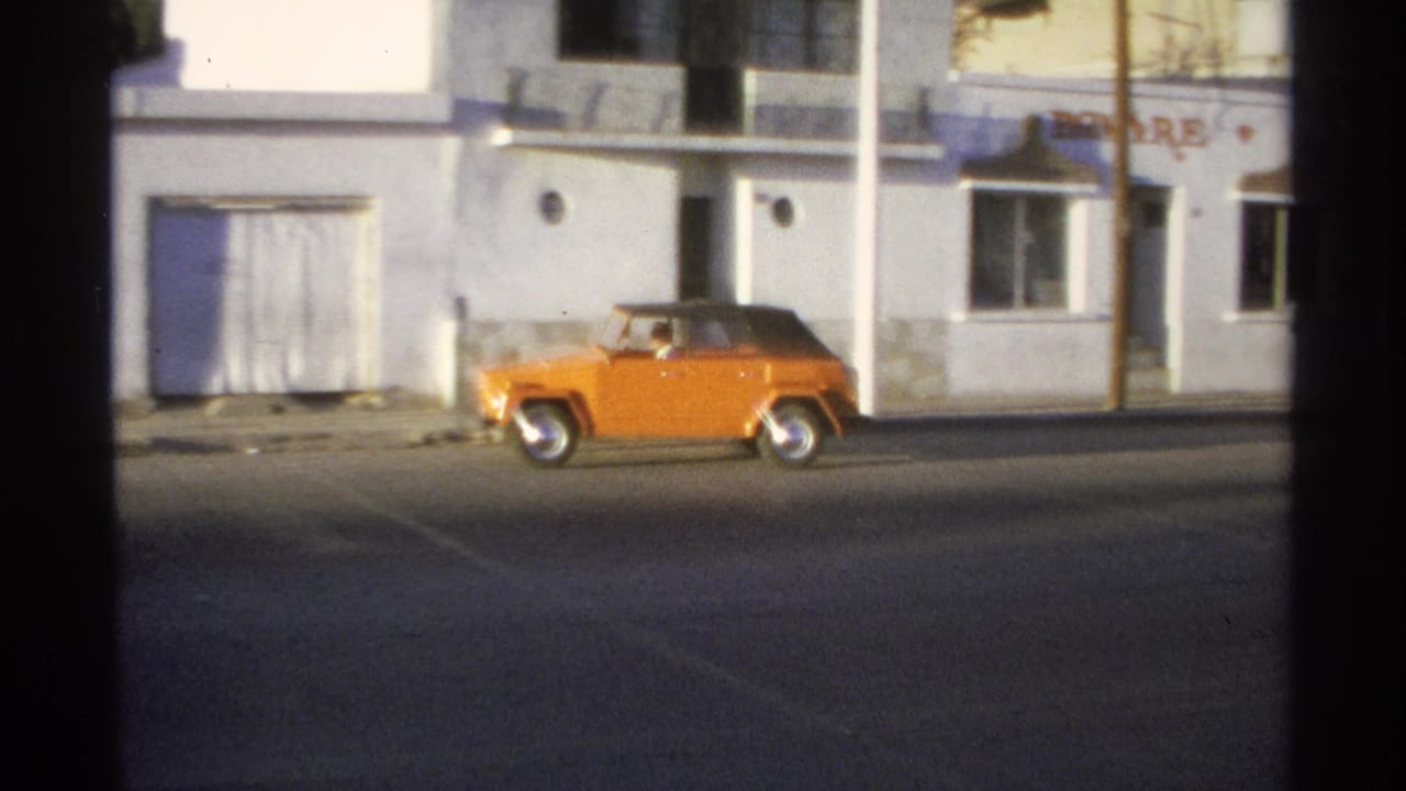 Orange soft-top car driving past white stucco buildings on a sunlit street.