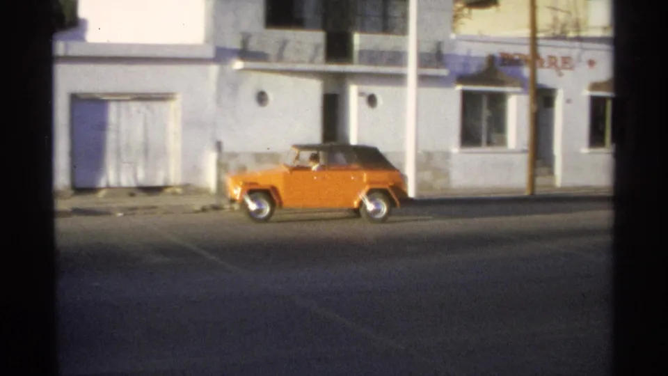 1975 Guaymas home-movie frame showing an orange Volkswagen Thing moving along a street past white stucco buildings.