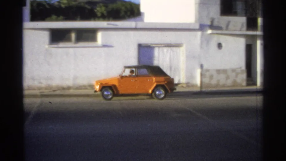 The orange Volkswagen Thing continues past a white stucco building in Guaymas, Mexico, with its dark soft top visible.