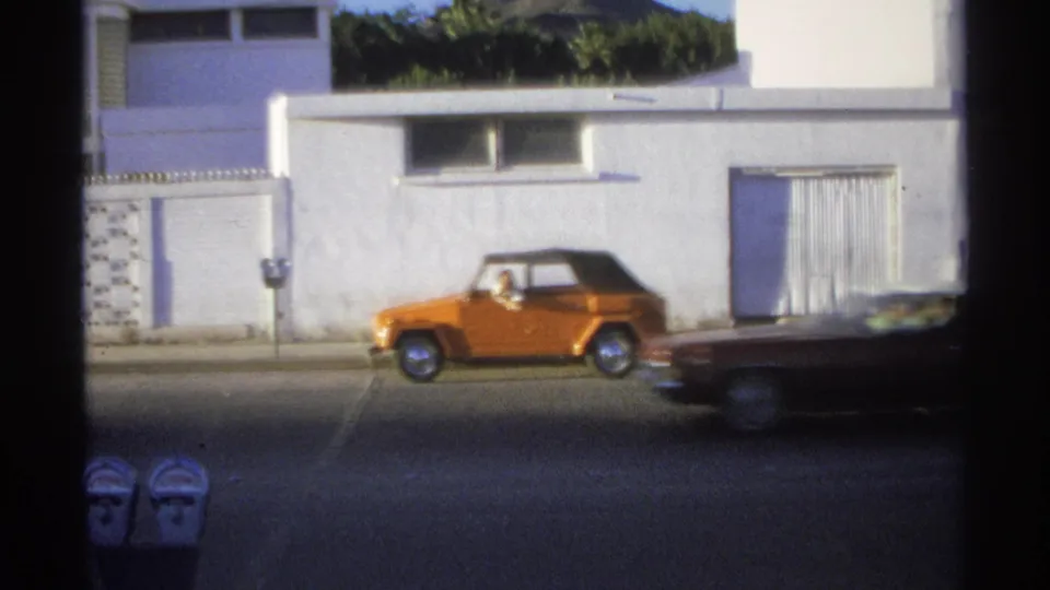 Guaymas street frame with the orange Volkswagen Thing near parking meters while a darker car crosses the foreground.