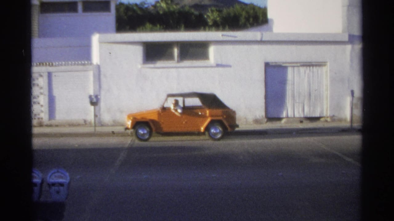 Orange convertible driving past a white building on a sunny street.