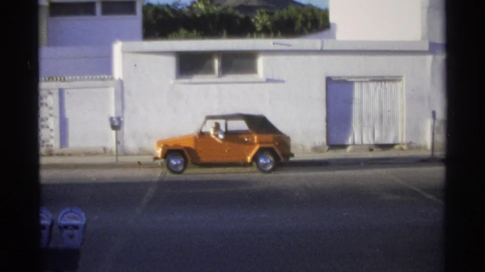 1975 Guaymas street view with an orange compact convertible passing a white building and shaded sidewalk.