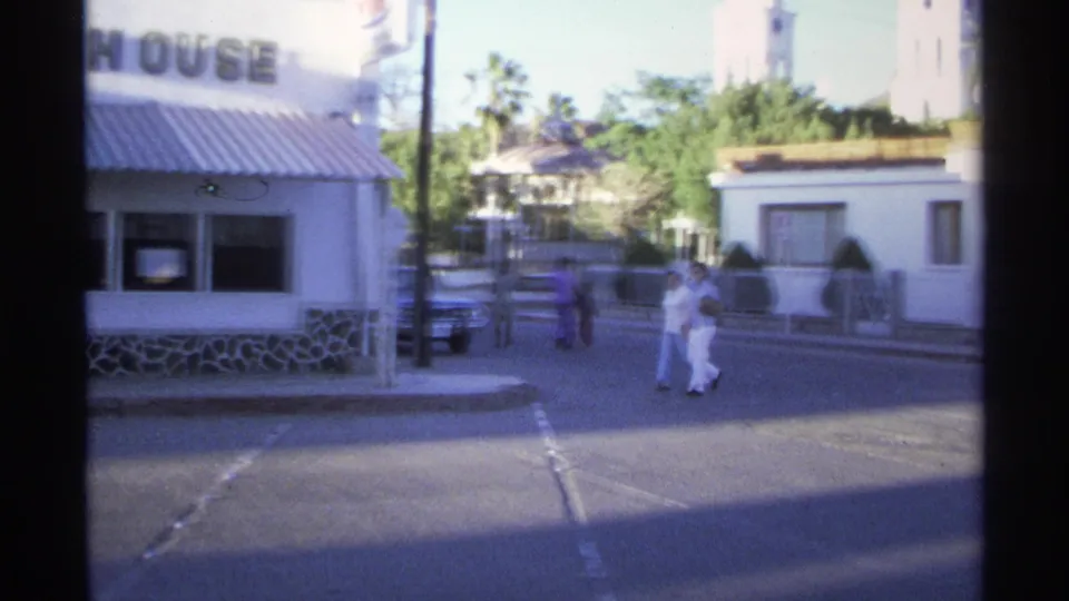 1975 Guaymas street scene with people crossing near a steak house, a parked car, palm trees, and white roadside buildings.