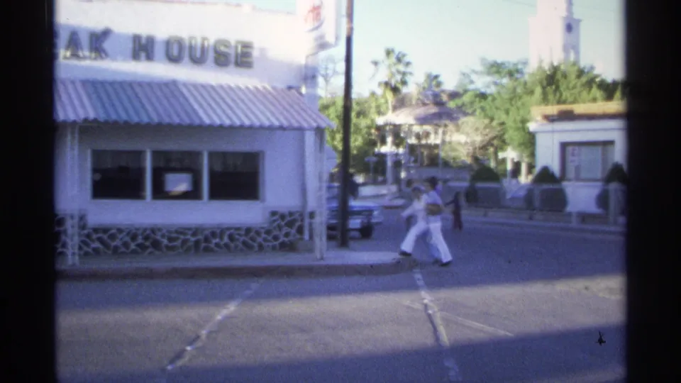 Guaymas street view with two pedestrians crossing by a steak house facade, parked car, white buildings, and palm trees.