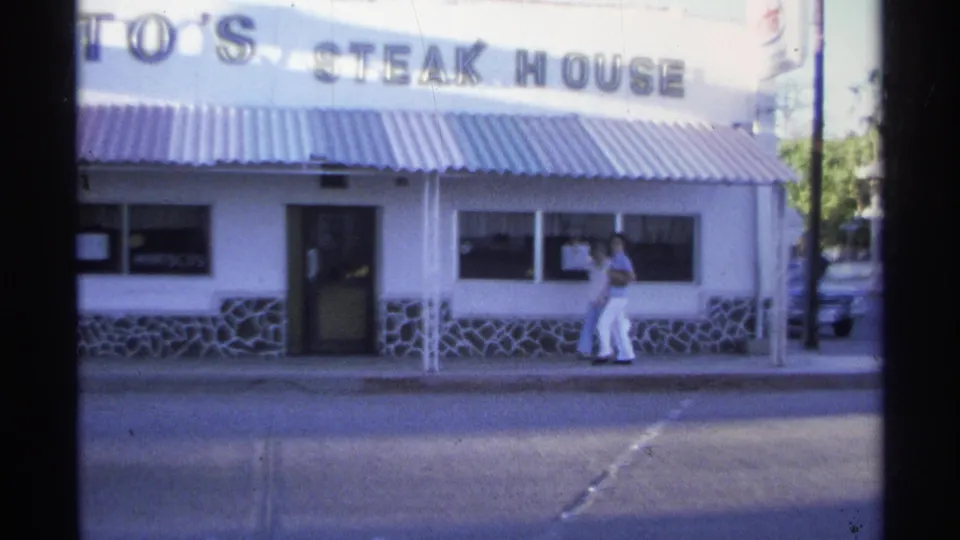 1975 Guaymas home-movie frame with a closer view of the steakhouse facade, including the door, windows, roof sign, and a person near the right-side window.