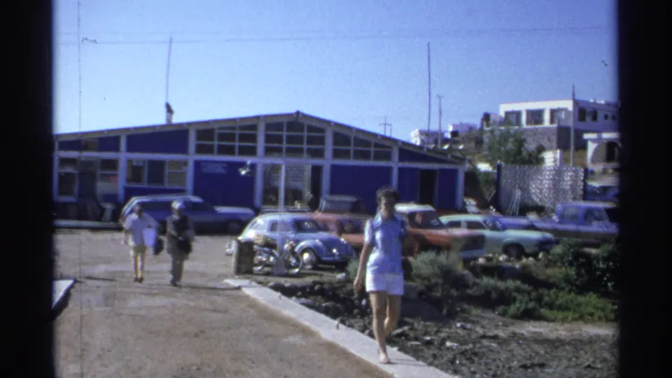 1975 Guaymas street-side frame showing a parking area with classic cars including a Volkswagen Beetle, pedestrians walking through the lot, and a low commercial building in bright sun.