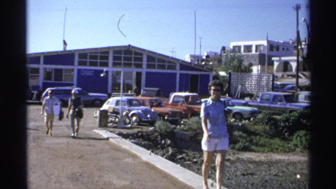 A woman in white shorts moves past the foreground in Guaymas while two pedestrians walk behind her near parked cars and a blue building.