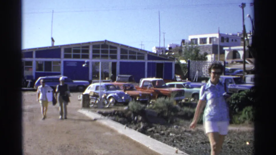 The foreground woman passes close to the camera, with a blue building, classic cars, and two walkers visible behind her in Guaymas.