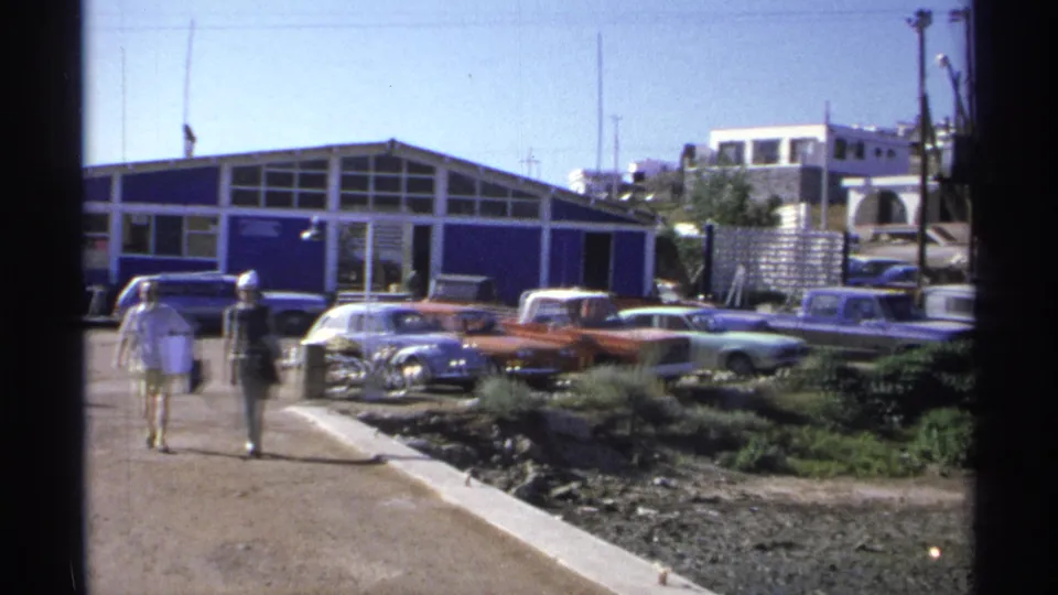 Two pedestrians continue along the path in Guaymas as the foreground walker leaves frame, with classic cars and a blue-and-white building behind them.